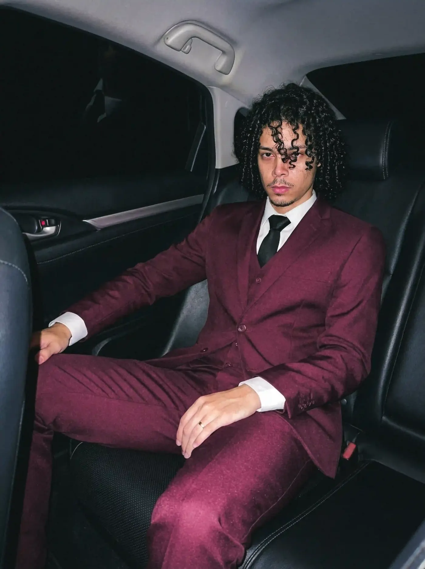 Bryan Haller vestindo terno vinho de três peças, olhar sério, no banco traseiro de um carro